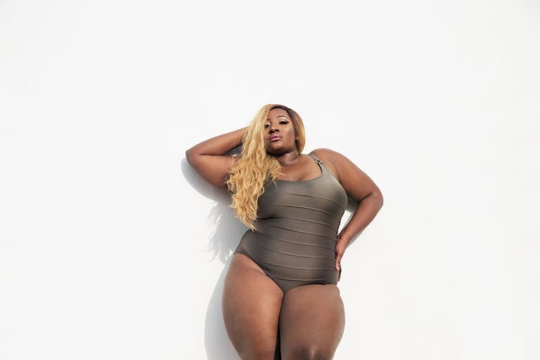 A confident black woman in a swimsuit posing against a white background, embracing body positivity and fashion.