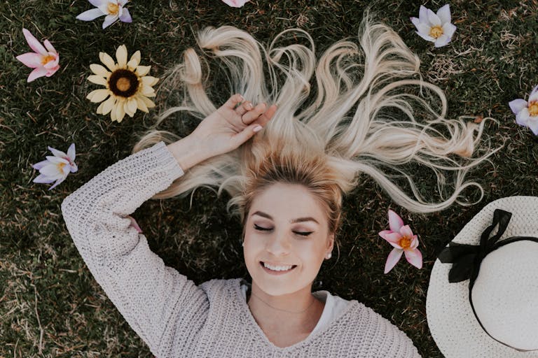 A smiling woman with closed eyes lies on grass, surrounded by flowers, capturing a moment of pure relaxation.