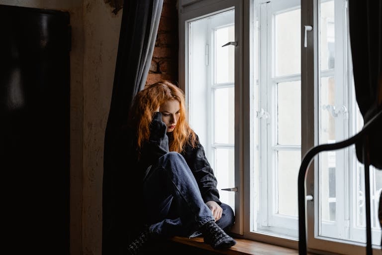 A woman sits by a window, lost in deep thoughts and moody light.