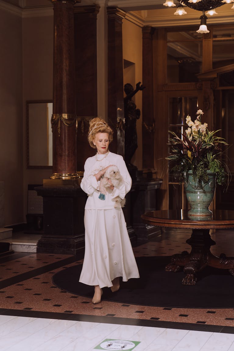 An elegantly dressed woman carrying a Maltese dog walks through a classic hotel lobby.