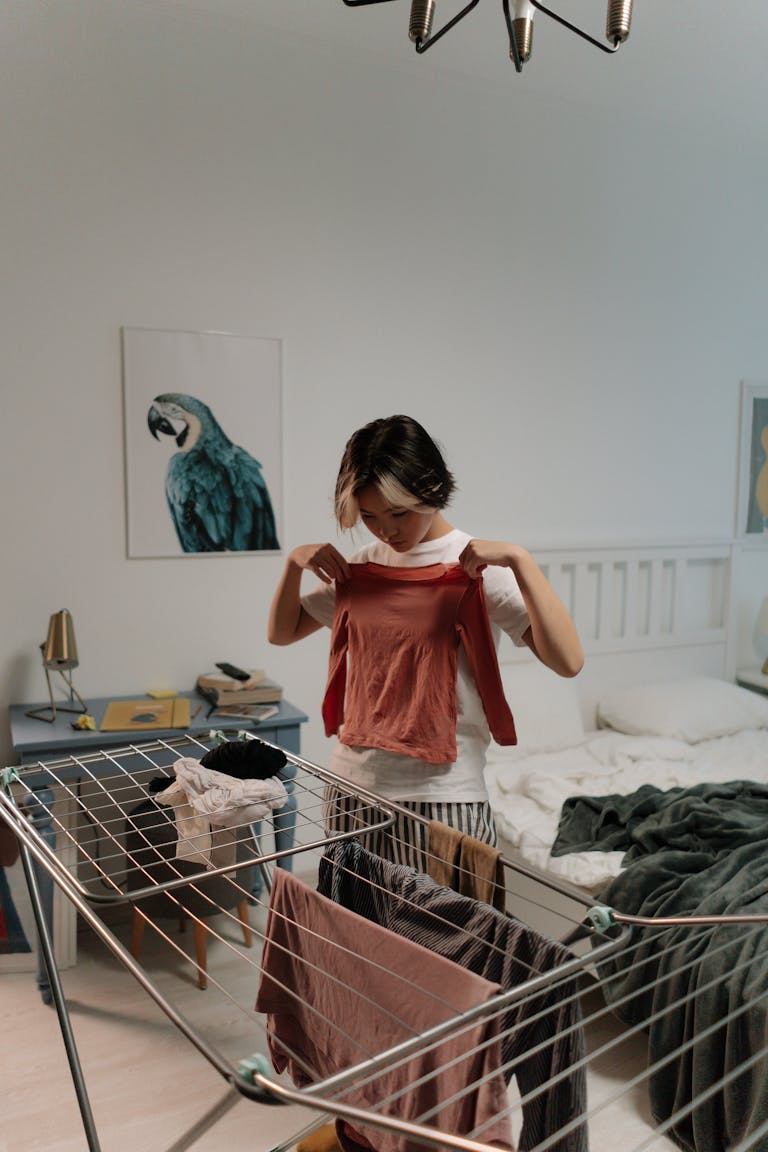 Asian woman folding clothes in a cozy home bedroom setting with a laundry rack.
