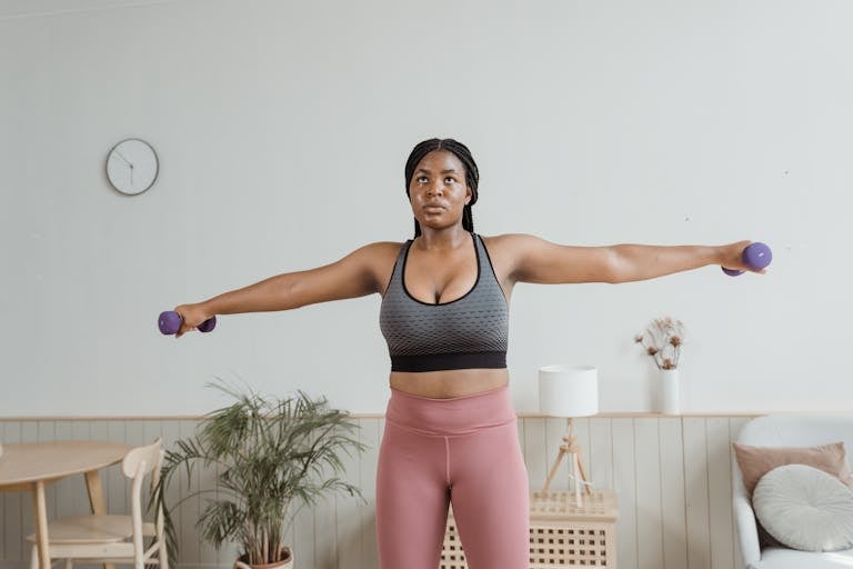 Black woman in activewear performing arm exercises indoors with dumbbells.