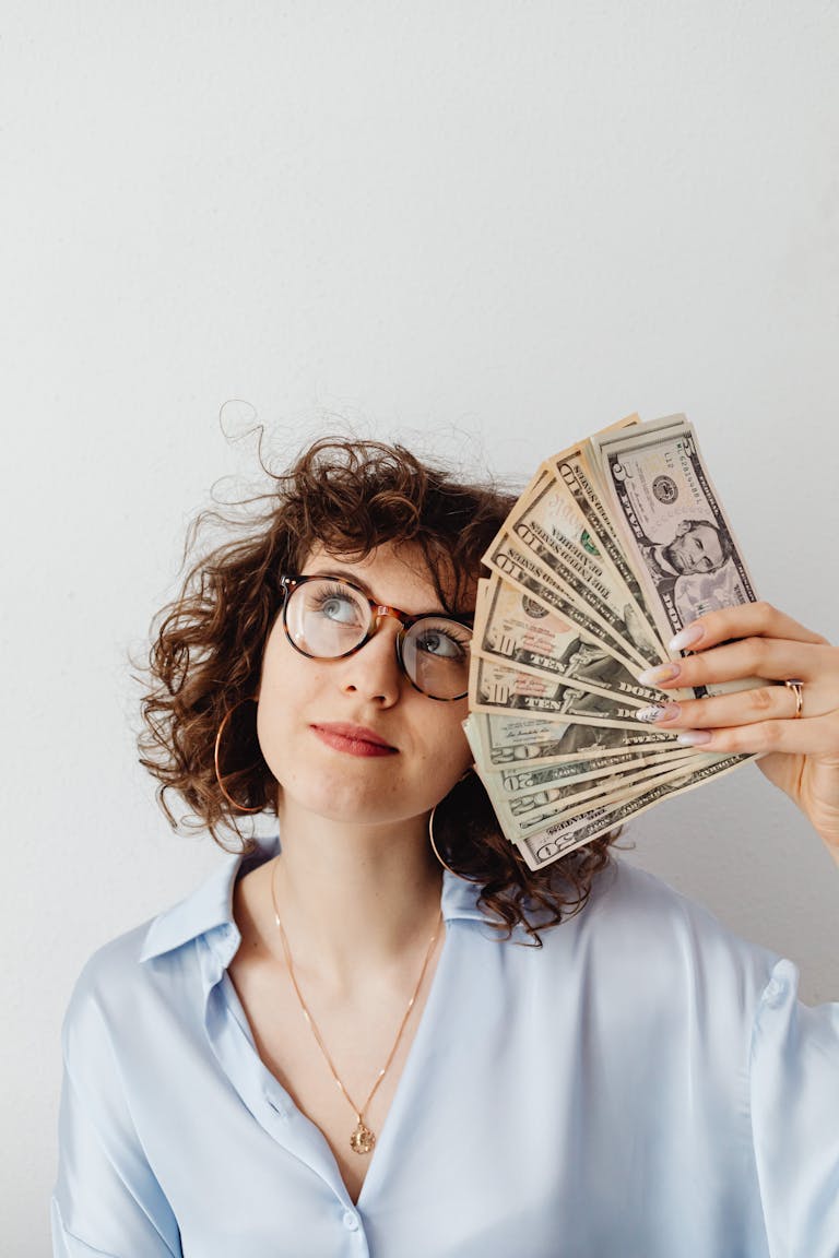 Caucasian woman happily holding a fan of US dollar bills against a light background.