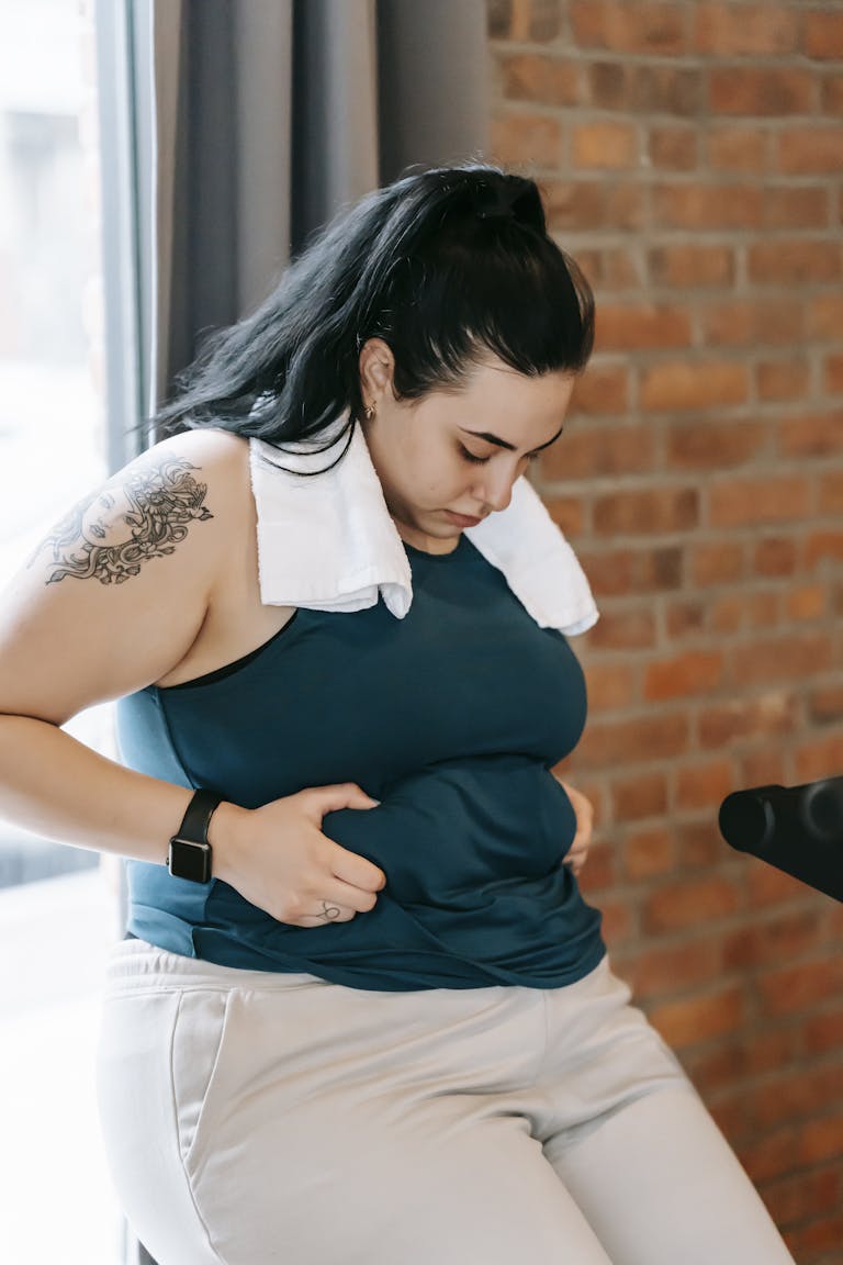 Upset young obese female in sportswear leaning on windowsill and desperately looking at stomach