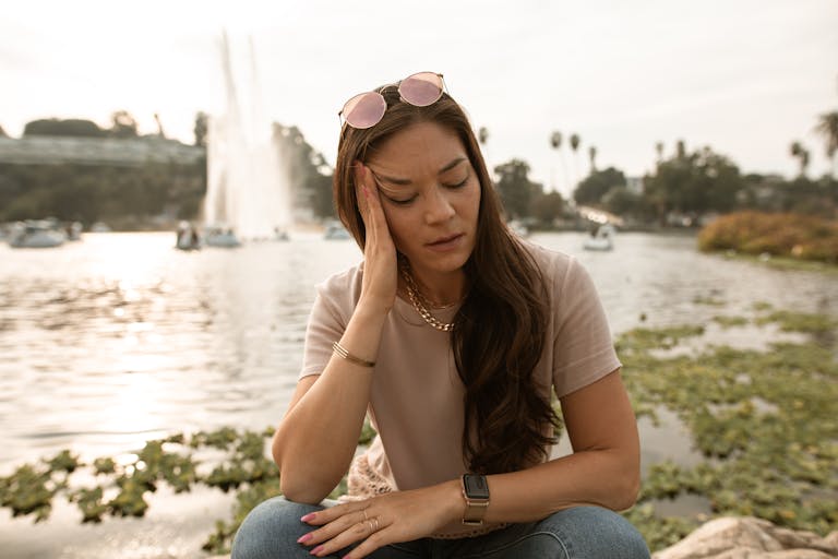 Woman feeling stressed and anxious, sitting by a lake with a serene background.