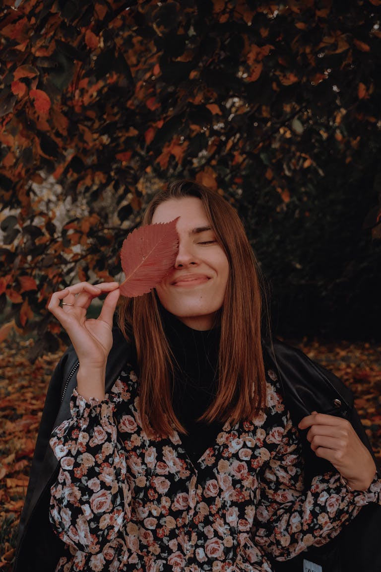 Woman in floral dress smiling with leaf over eye in autumn setting.